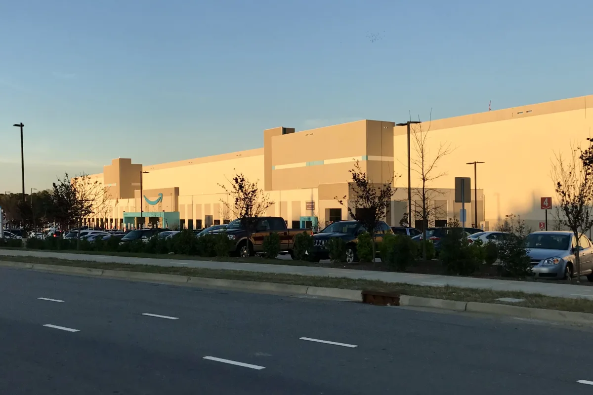 A large beige Amazon fulfilment centre beside a road with parked cars