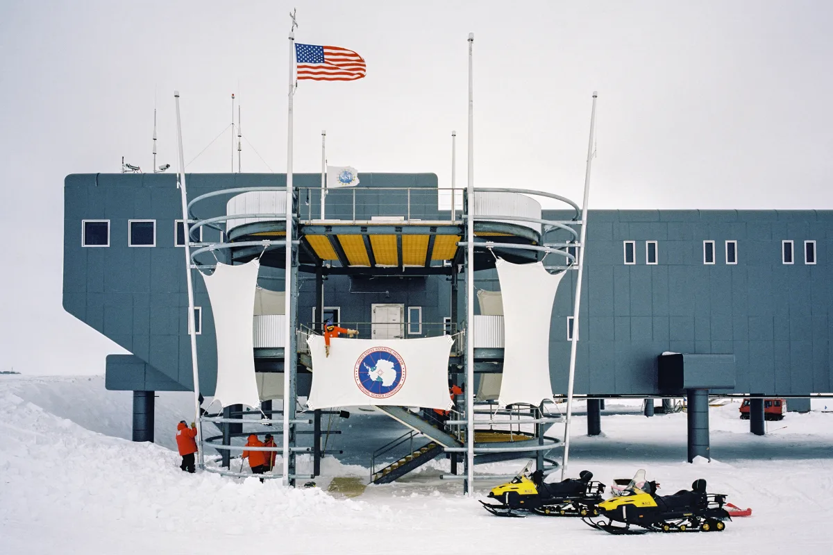 The raised blue-grey Amundsen-Scott South Pole Station building on the snow with flags and snowmobiles
