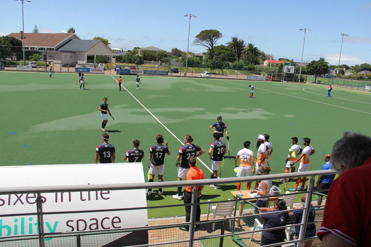 Hockey players on an artificial pitch seen from the stands, with substitutes along the touchline